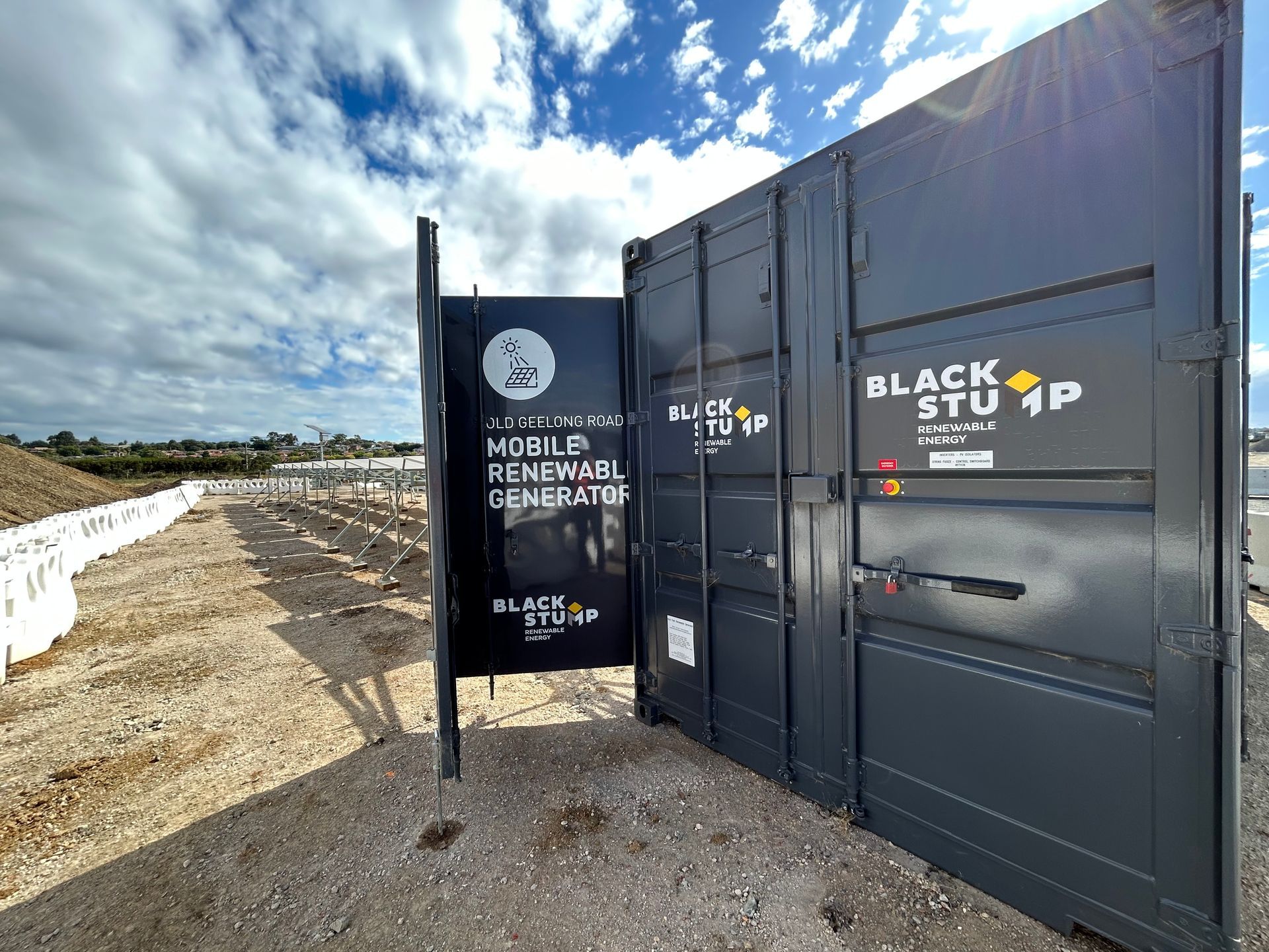 Black, industrial container with open door; solar panel field in the background under cloudy sky.