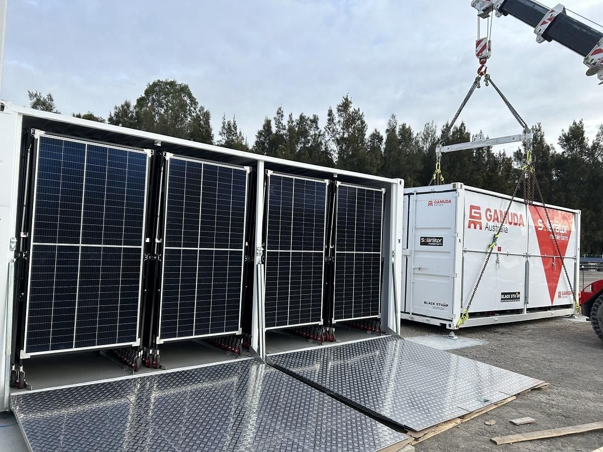 Crane lifting a container from a solar panel array container outdoors. Blue sky, grey metal, and red and white branding are visible.