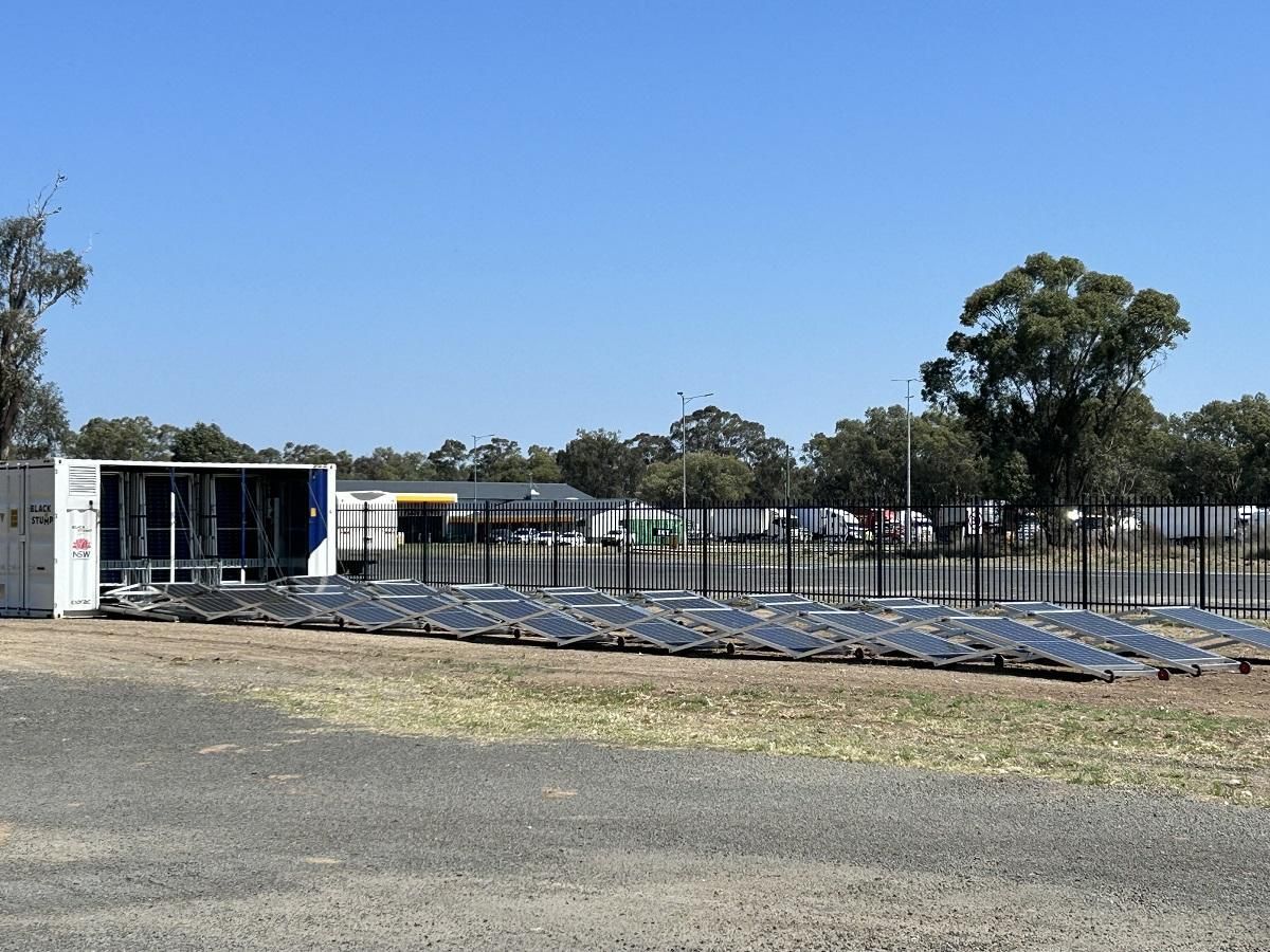 Solar panels laid out next to a shipping container, in front of a fence and a building under a blue sky.