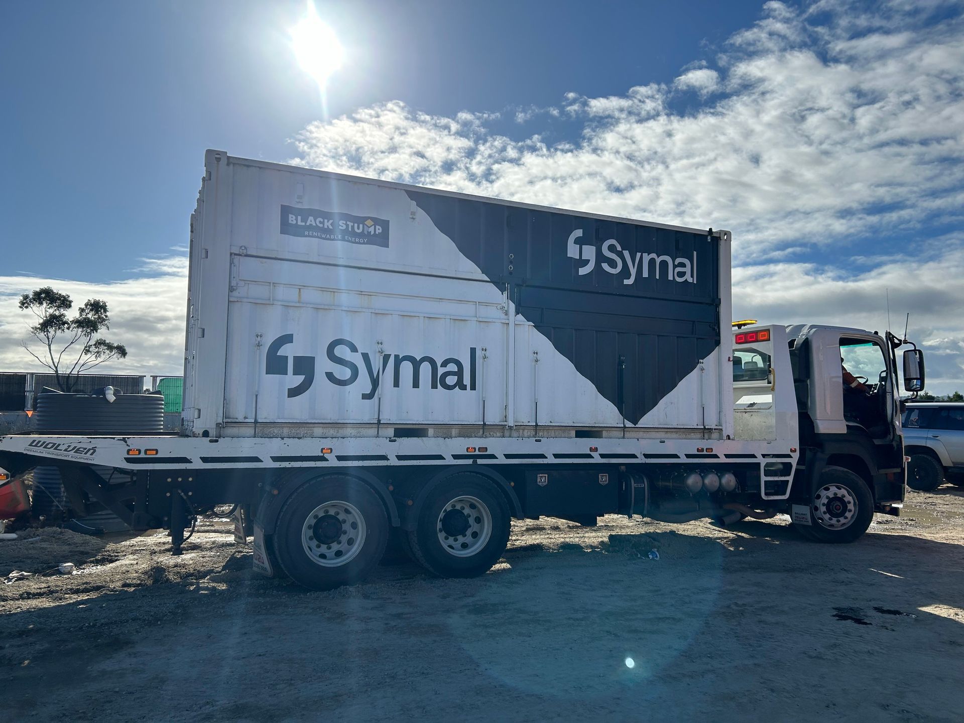 Truck carrying a large container labeled "Symal" on a flatbed under a bright, sunny sky.