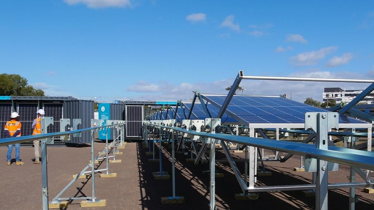 Solar panels on a rooftop, supported by metal frames, with two workers nearby under a blue sky.