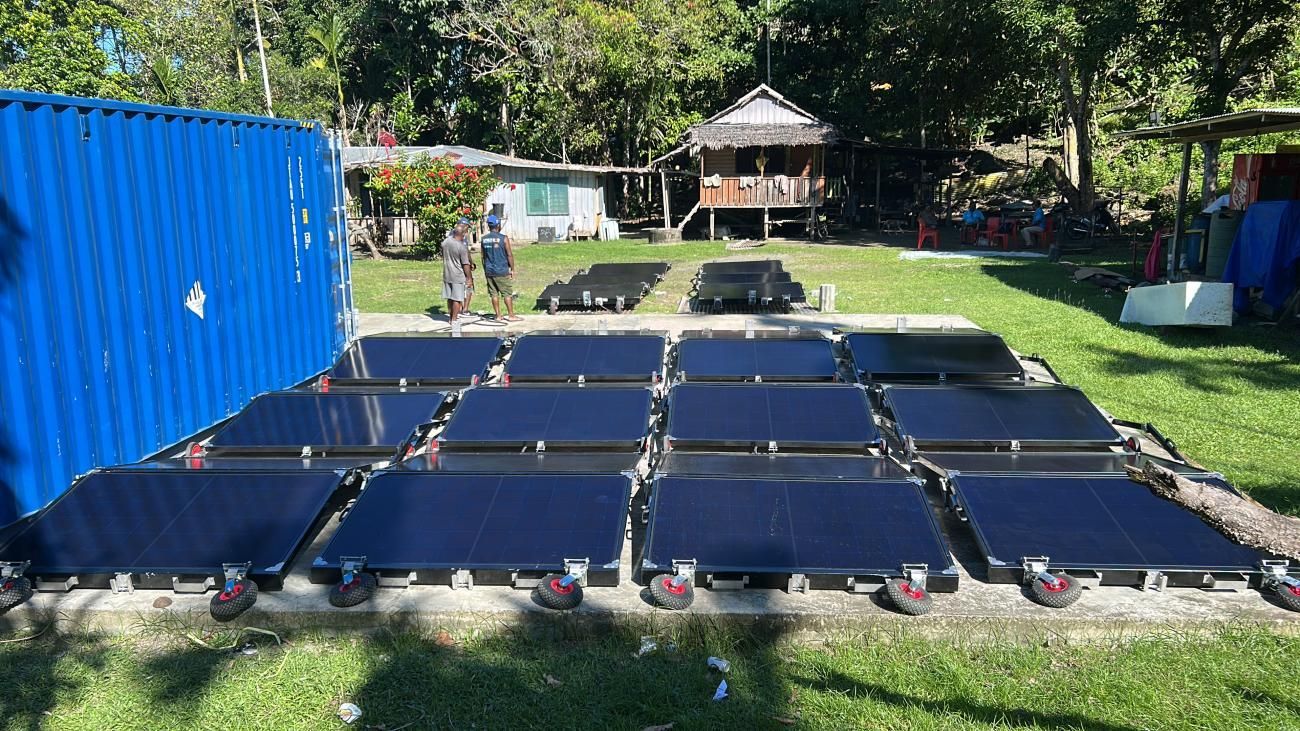 Solar panels laid out on grass next to a blue container and small buildings.