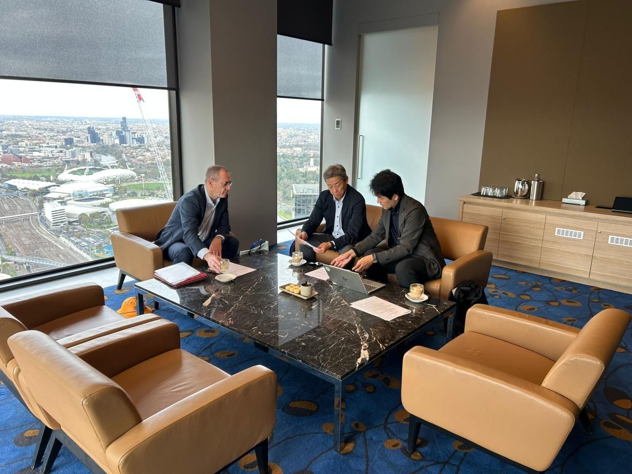 Three men in suits seated around a table, discussing documents in a high-rise office with city views.
