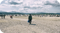 Man walks away from a refugee camp with tents. Mountains and cloudy sky in background.