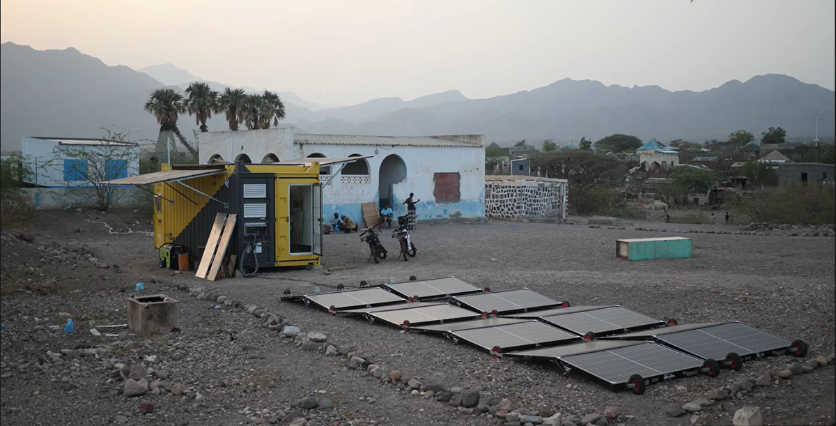 Solar panels with people setting up near a yellow structure and buildings in a mountainous landscape.