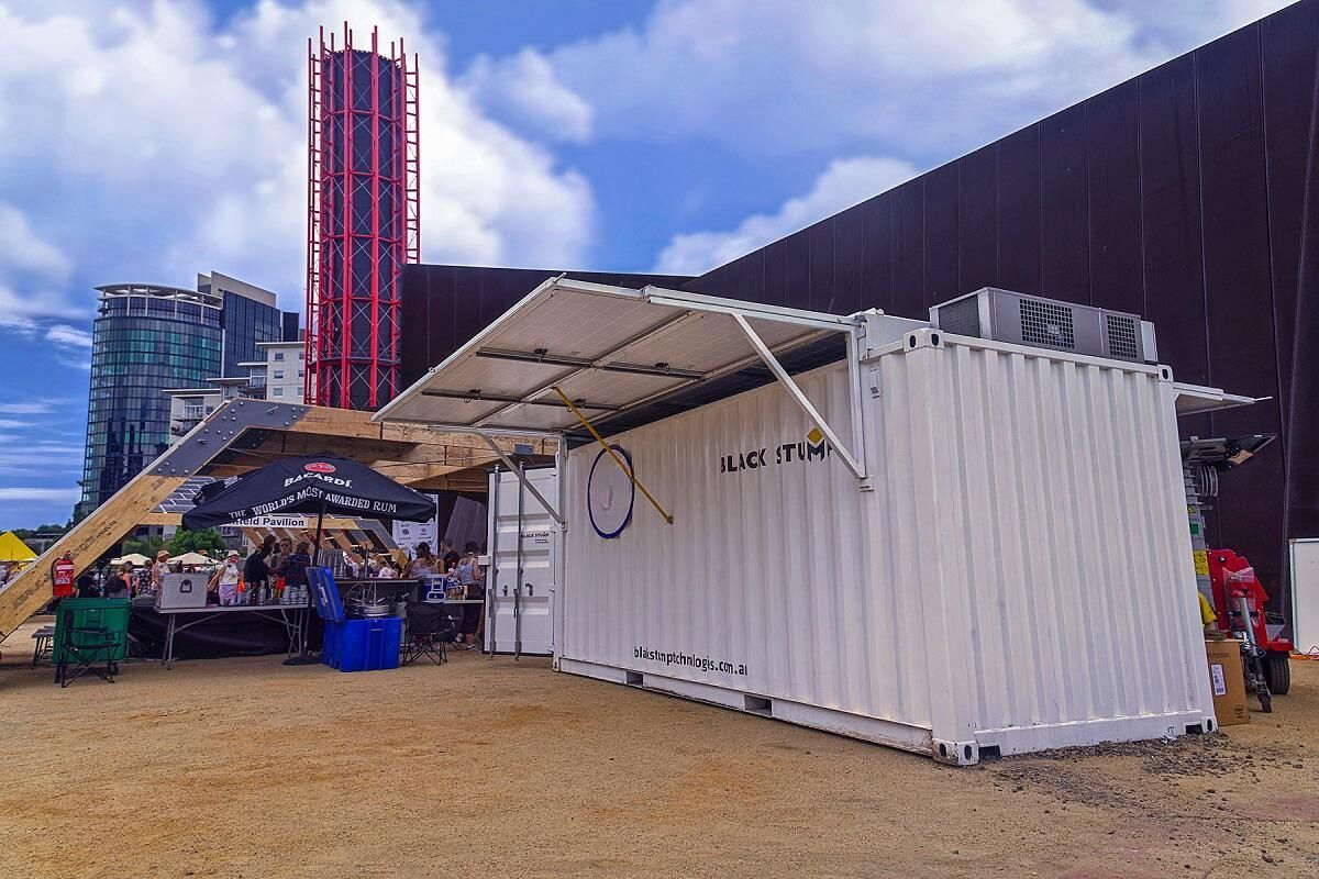White shipping container converted into a food/drink stand with a canopy. People gathered outside on a sunny day.
