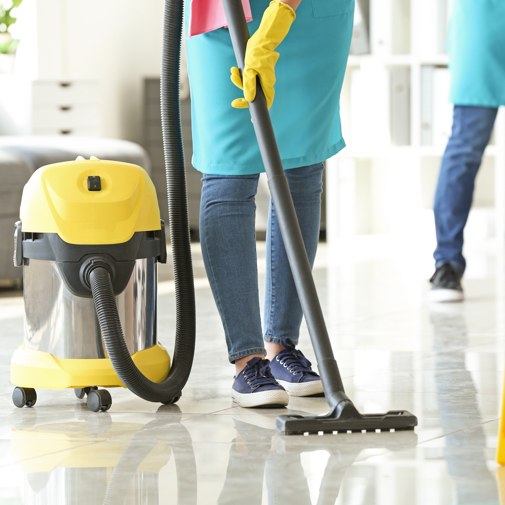 Person in blue apron, yellow gloves vacuums a shiny floor, another person in background.