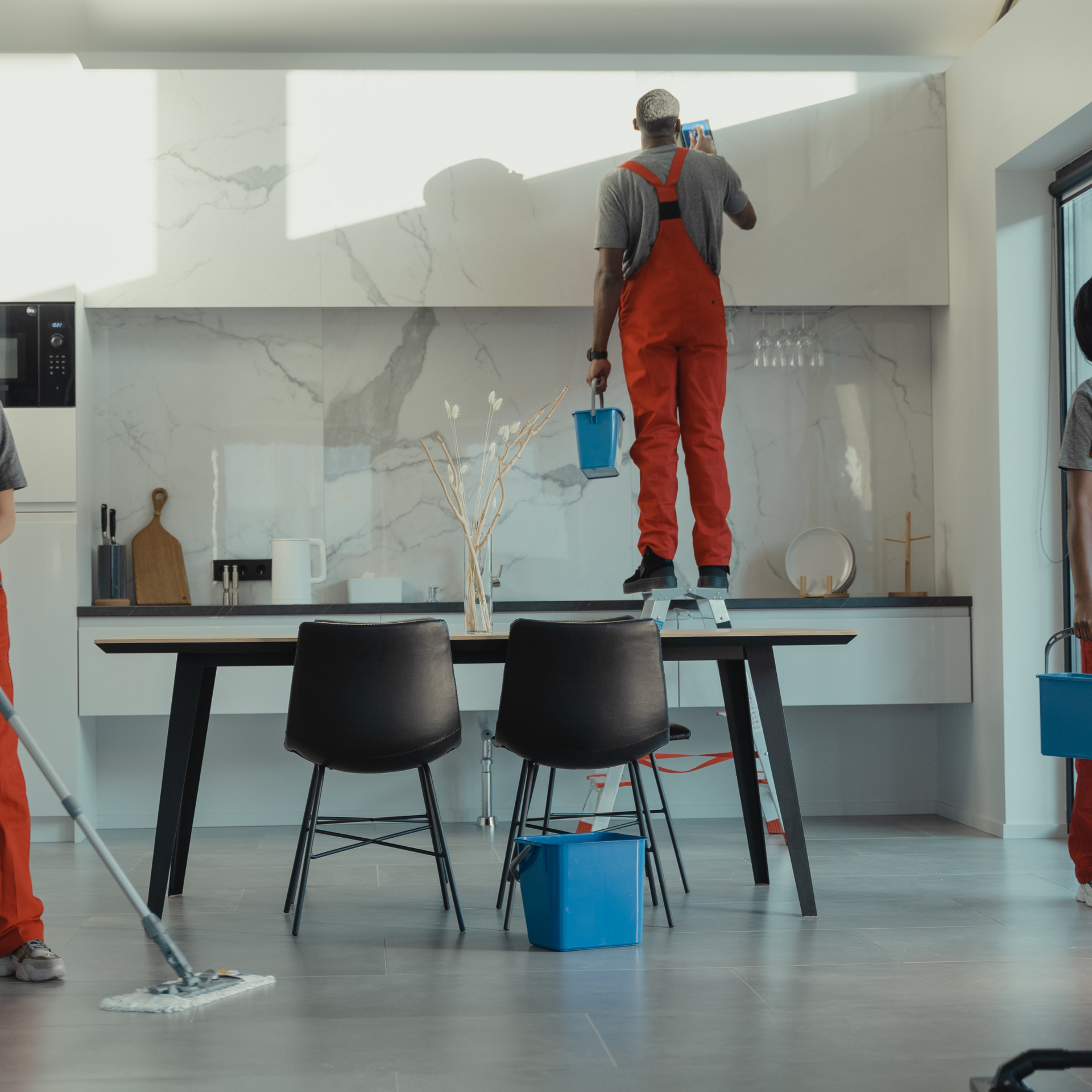 Three people cleaning a modern kitchen. One on a table, washing wall, others mopping and holding buckets.