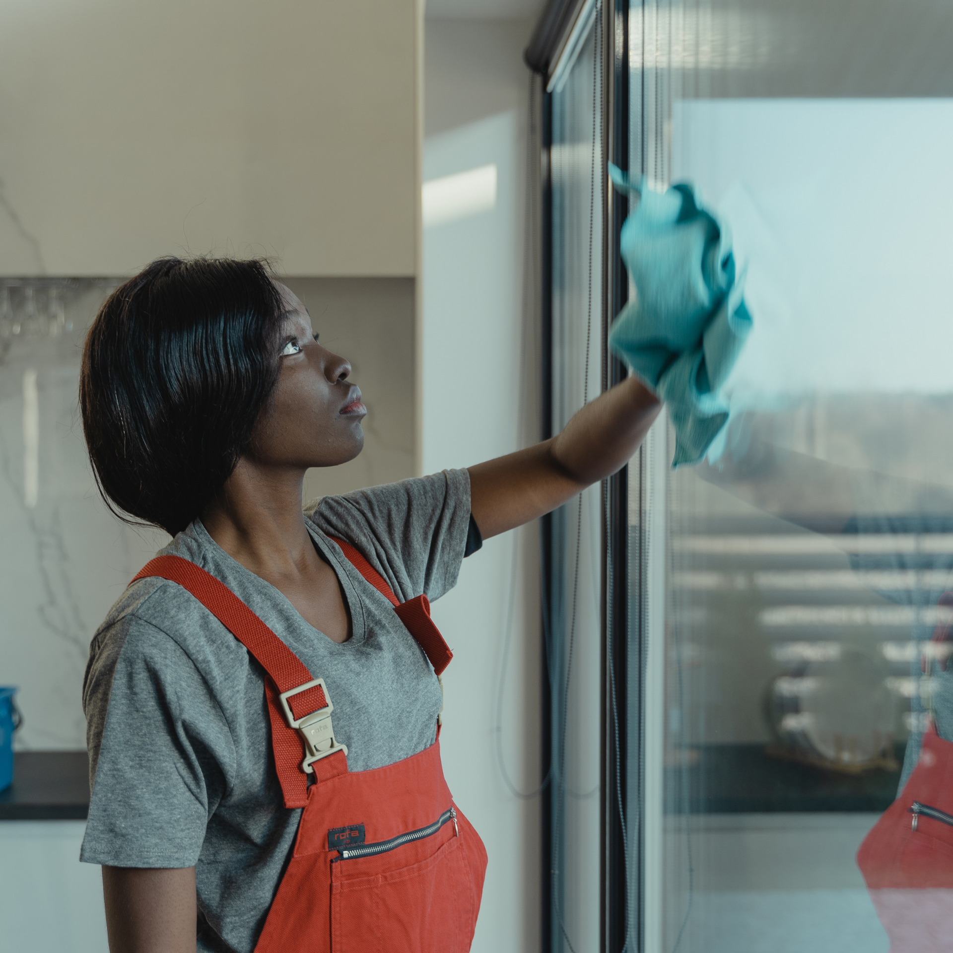 Woman in red overalls cleaning a window with a blue cloth.