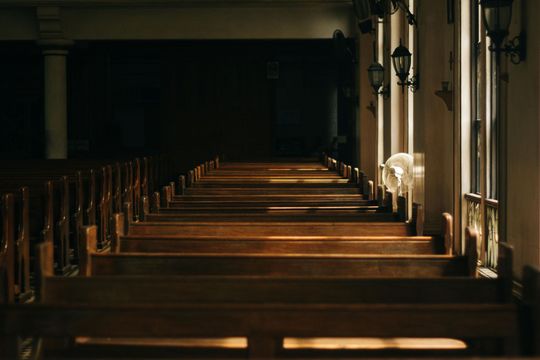 Rows of wooden church pews lead to a dark altar. Light streams through a window on the right.