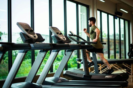 Man running on a treadmill in a gym with large windows.