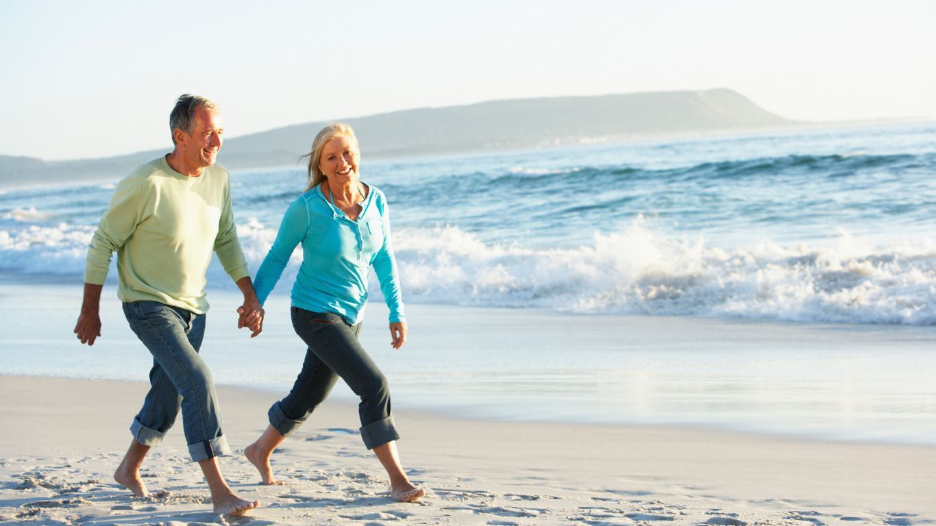 An elderly couple is walking on the beach holding hands.