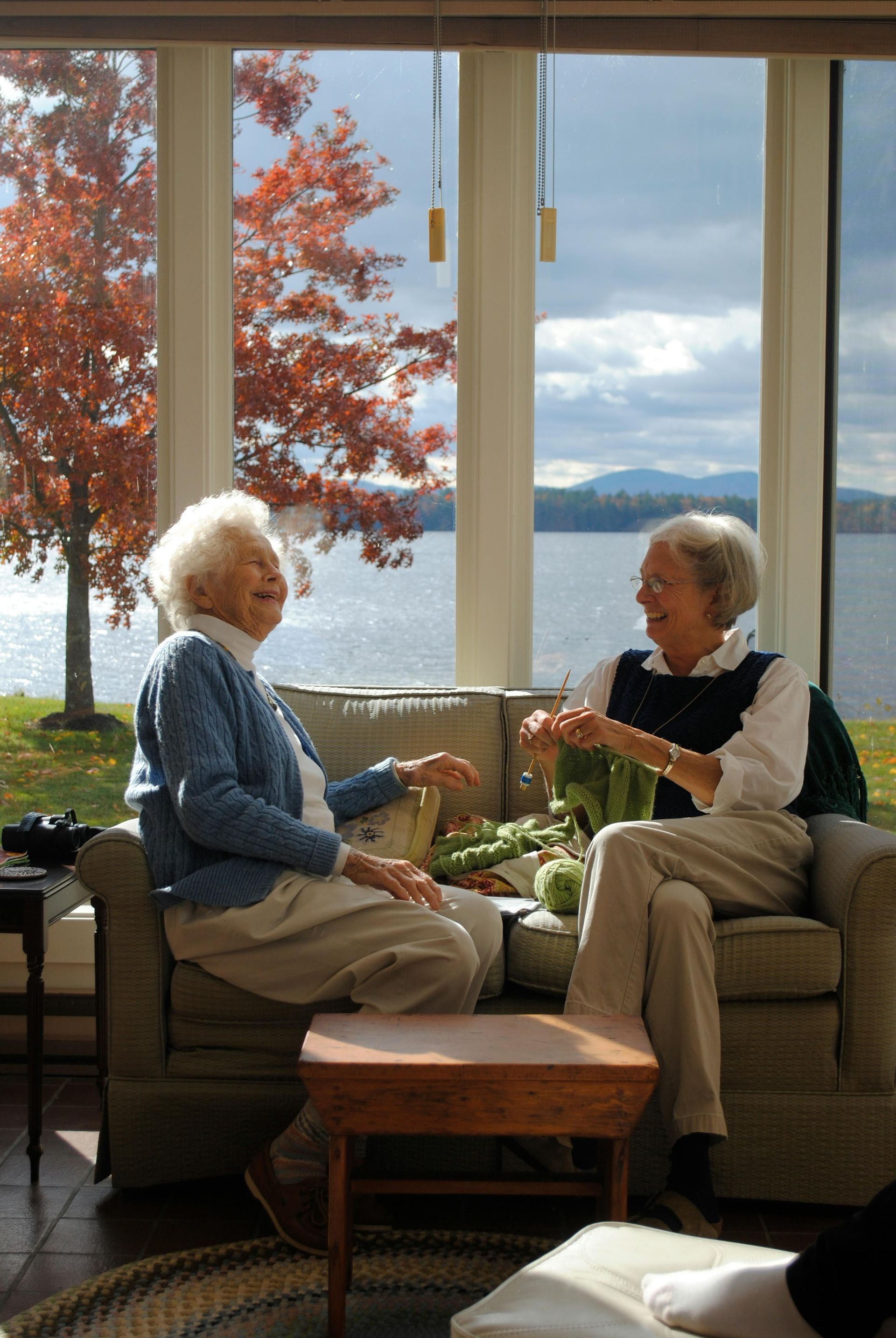 Two elderly women are sitting on a couch in front of a window overlooking a lake.