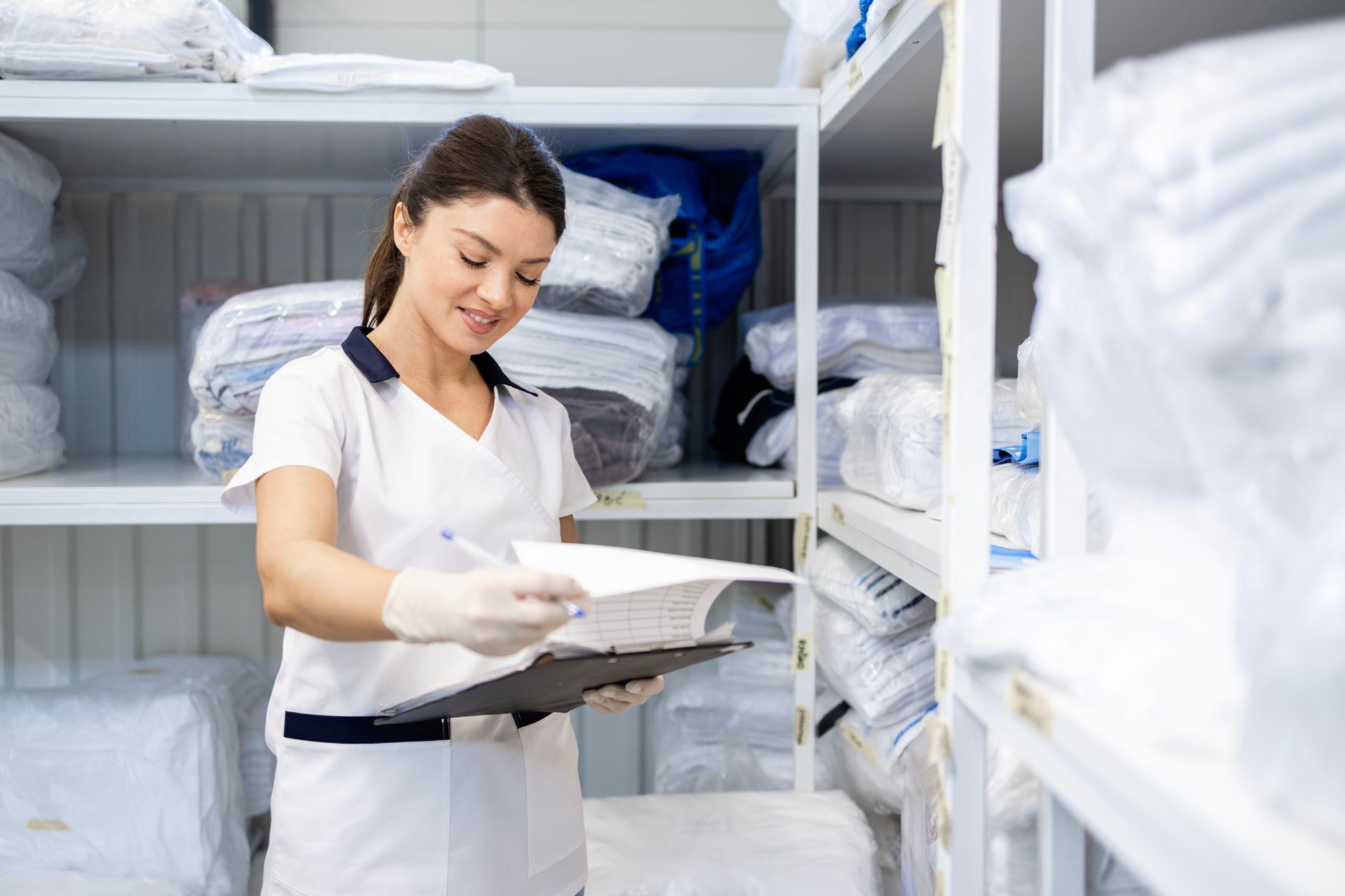 Woman in white uniform with clipboard checks inventory in a storage room filled with linens.