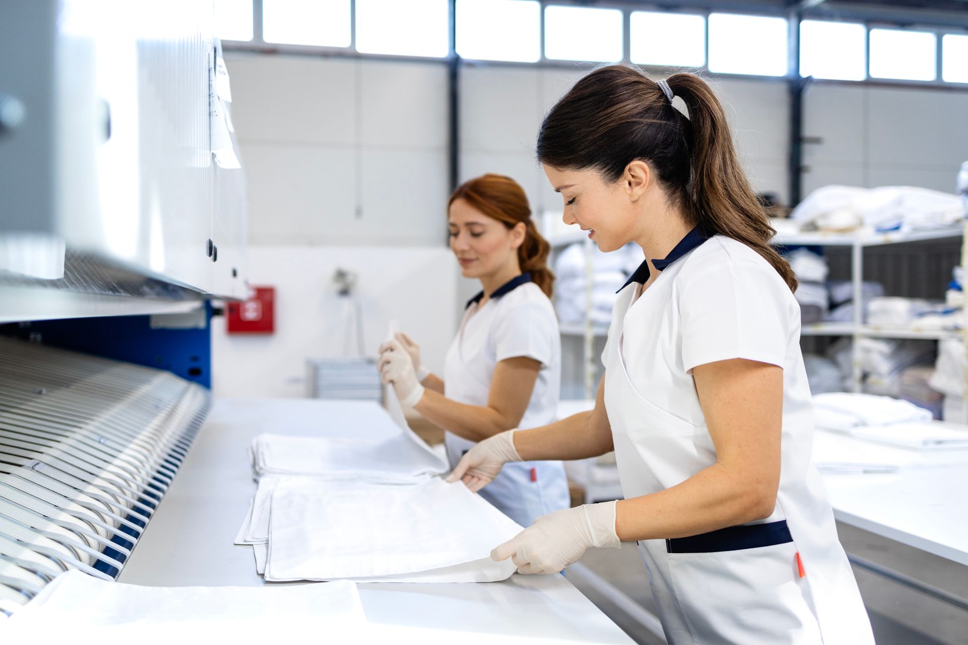Two women folding white linens in a commercial laundry facility, wearing white uniforms and gloves.