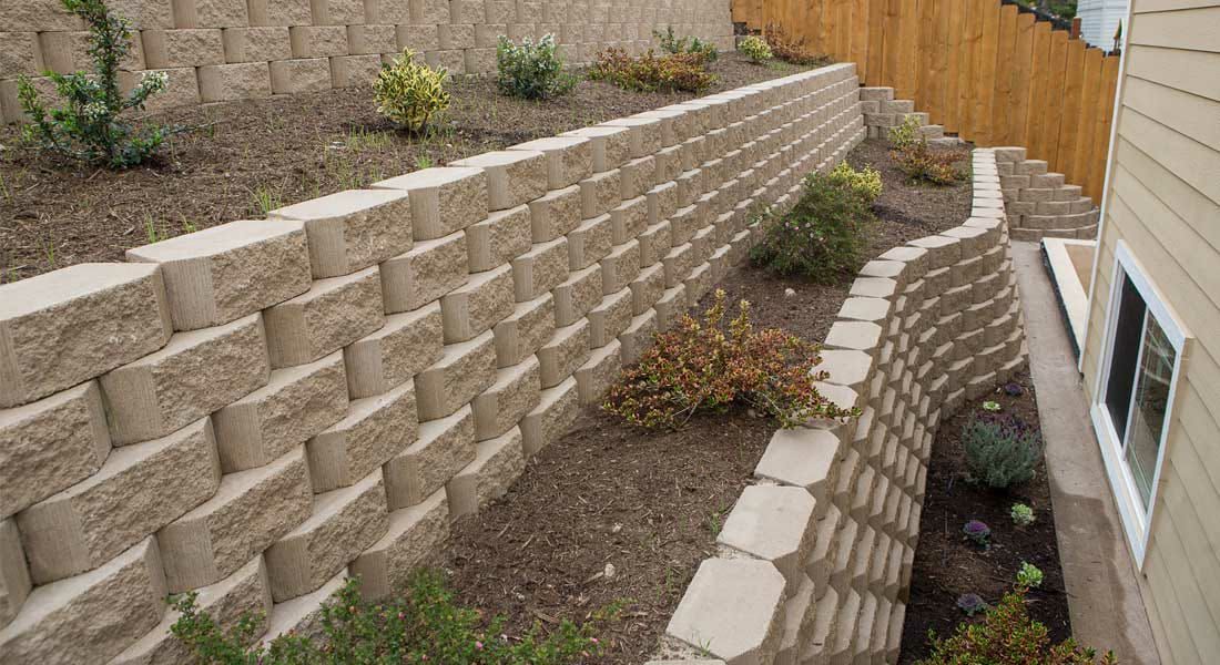 A brick retaining wall is surrounded by plants and a wooden fence.