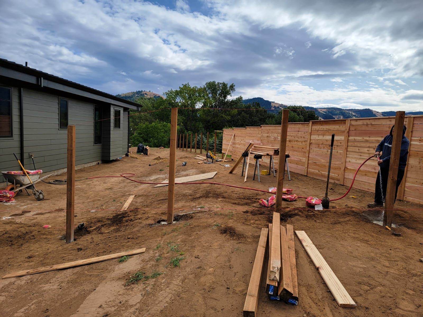 Fence construction: wooden posts set in dirt yard, near a house and person using a tool.