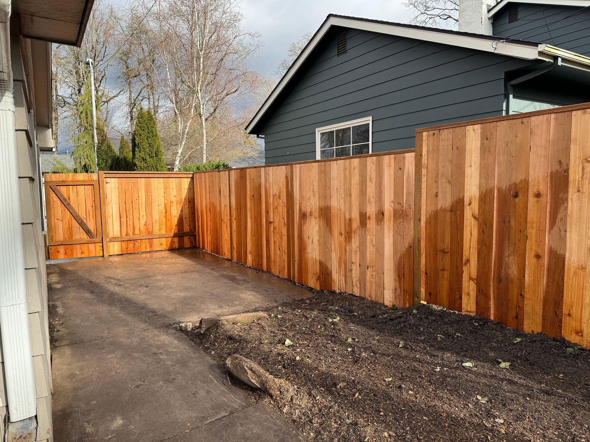 Wooden fence bordering a driveway, with a gate, next to a dark-colored house. Brown soil in the foreground.