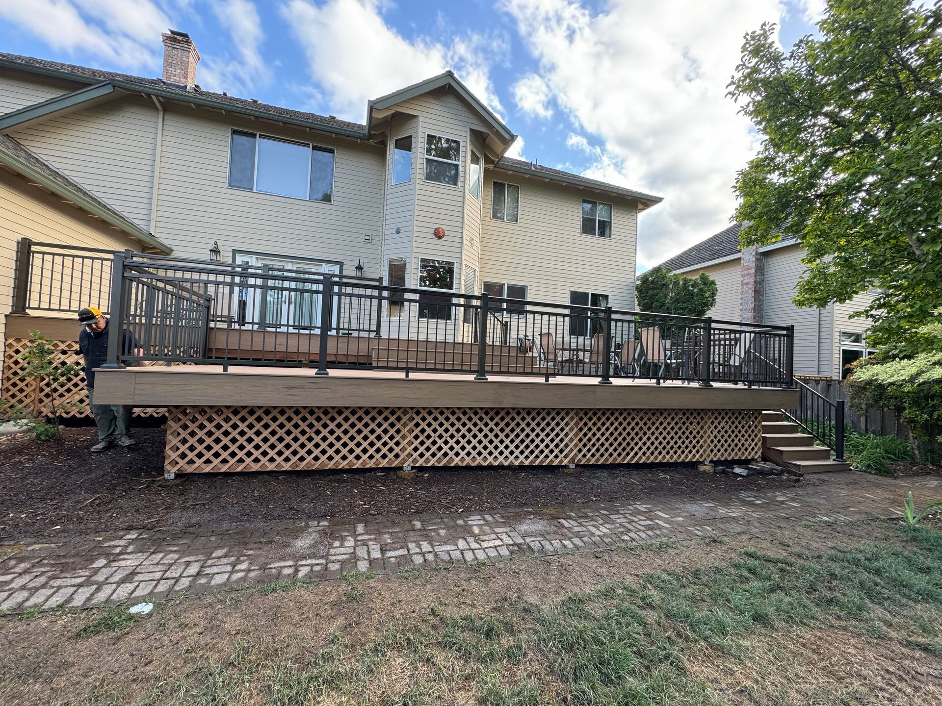 Rear view of a house with a large wooden deck. Dark railing, tan siding, and lattice work on the deck.