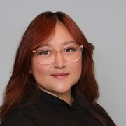 Portrait of a person with red hair and clear glasses, wearing a black top against a gray background