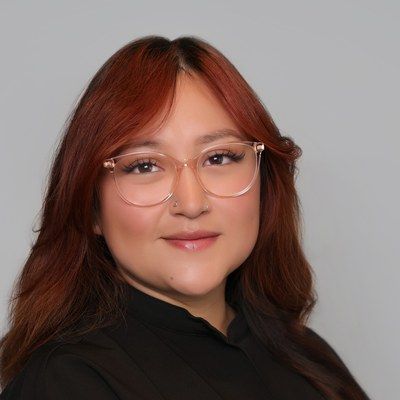 Portrait of a person with red hair and clear glasses, wearing a black top against a gray background