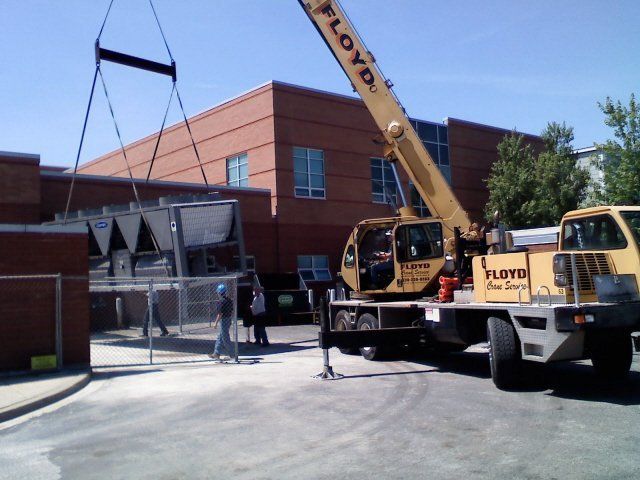 A crane lifts HVAC equipment over a building. Yellow crane, people, and a sunny outdoor setting.