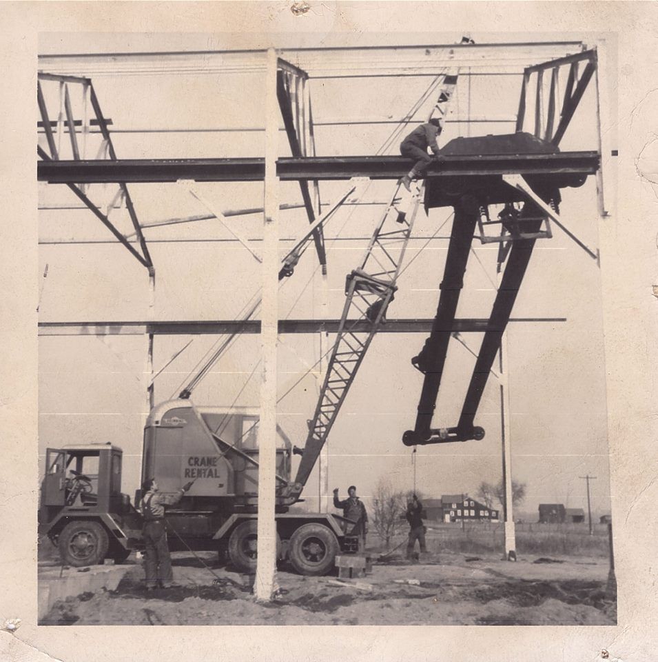 Construction workers using a crane to lift steel beams at a building site. Crane rental truck present.