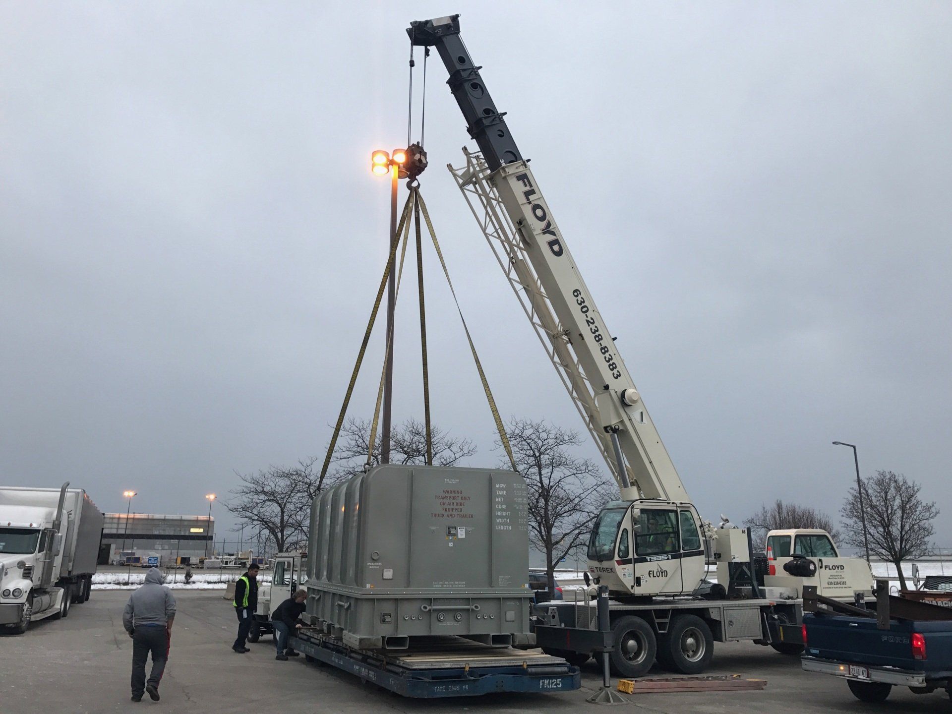 A crane lifting a large gray container onto a flatbed truck in a parking lot on an overcast day.