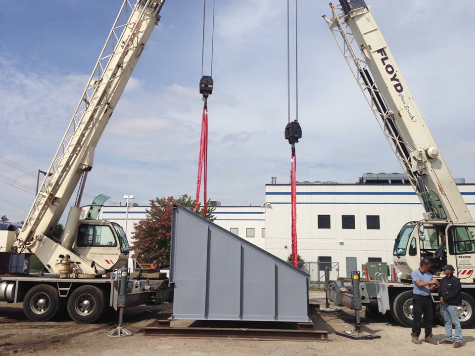 Two cranes lifting a large, gray, angled structure outdoors on a sunny day. Two people watch.