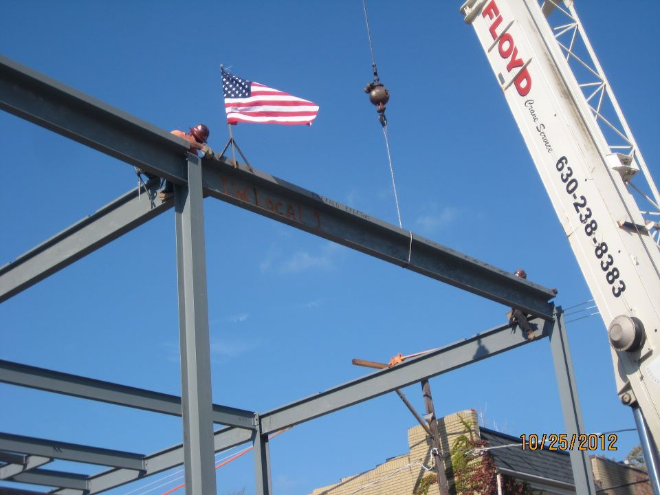 Steel Erectors — Structure Erectors in Wood Dale, IL Construction workers raising a steel beam with a crane, American flag on the structure, clear blue sky.