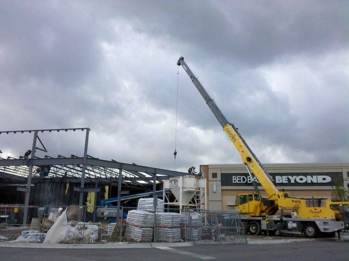 Construction site with crane lifting steel beams; a Bed Bath & Beyond store is visible in the background under a cloudy sky.