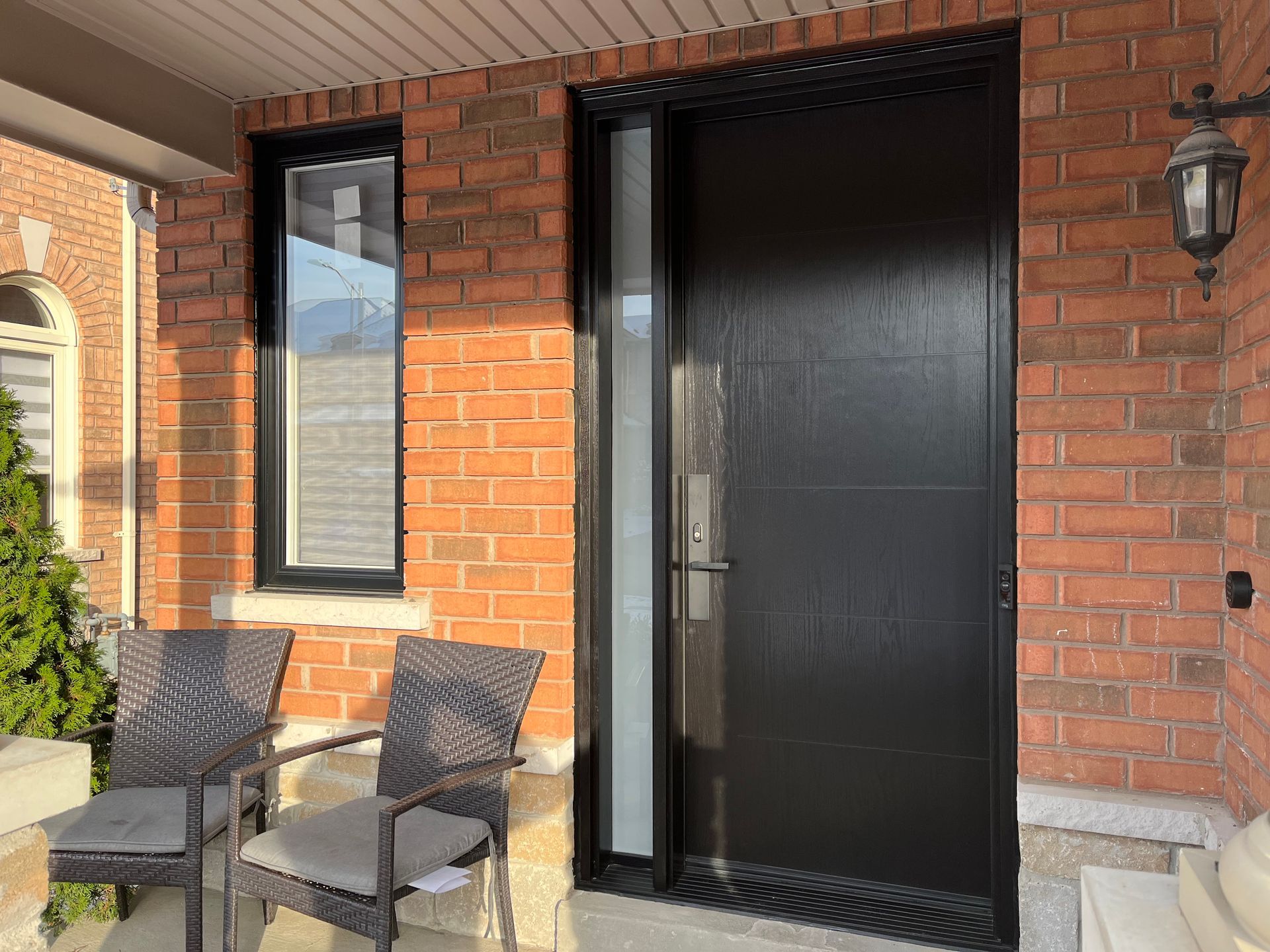 The front door of a brick house with a black door and wicker chairs.