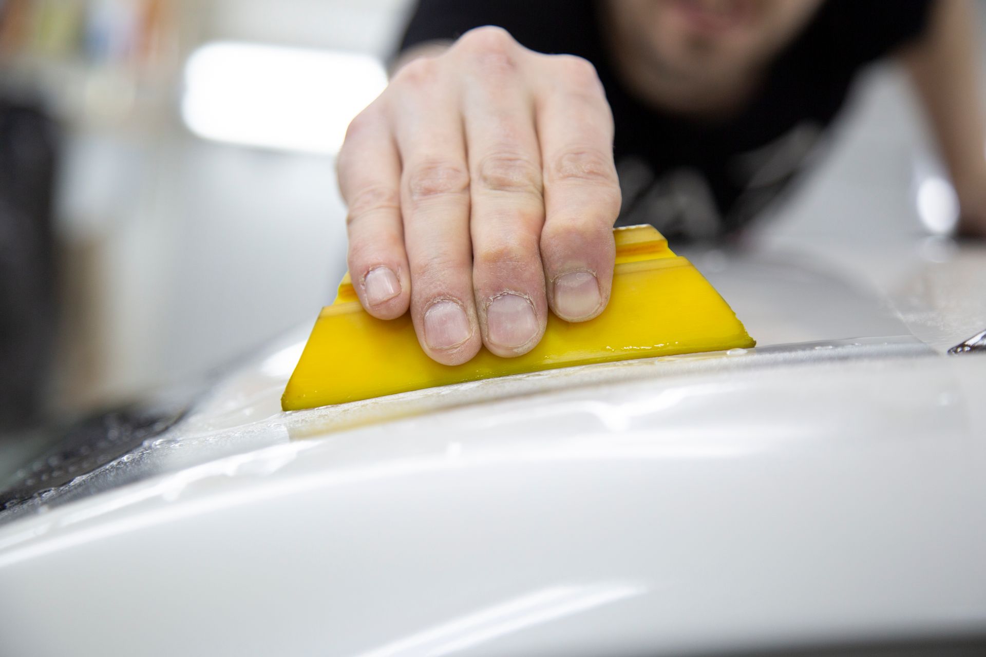 Mano di una persona che usa una spatola gialla per applicare una pellicola sulla superficie bianca di un'auto.
