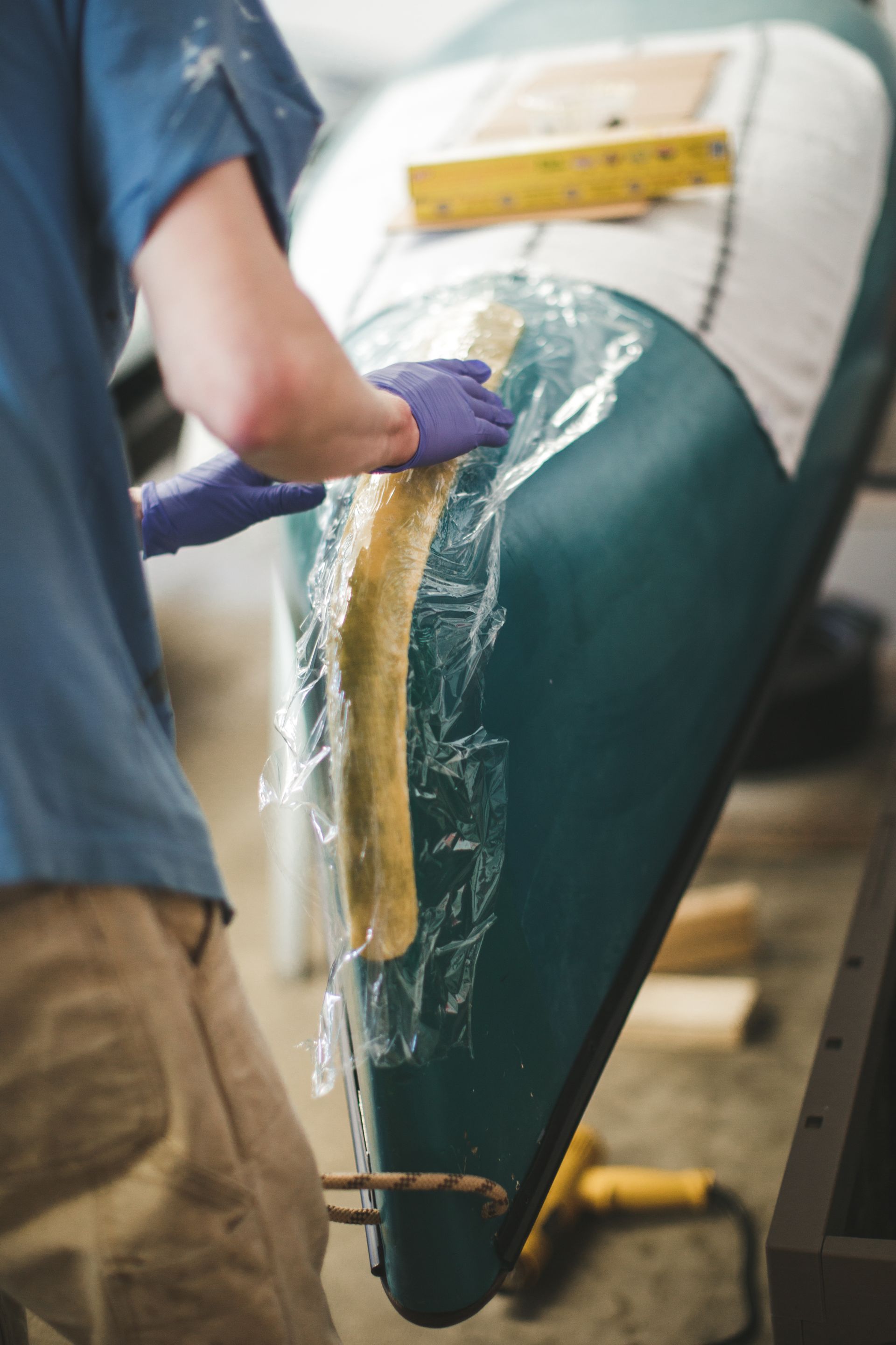 Una persona con una maglietta blu ripara lo scafo di una canoa verde con pellicola trasparente e mani guantate.
