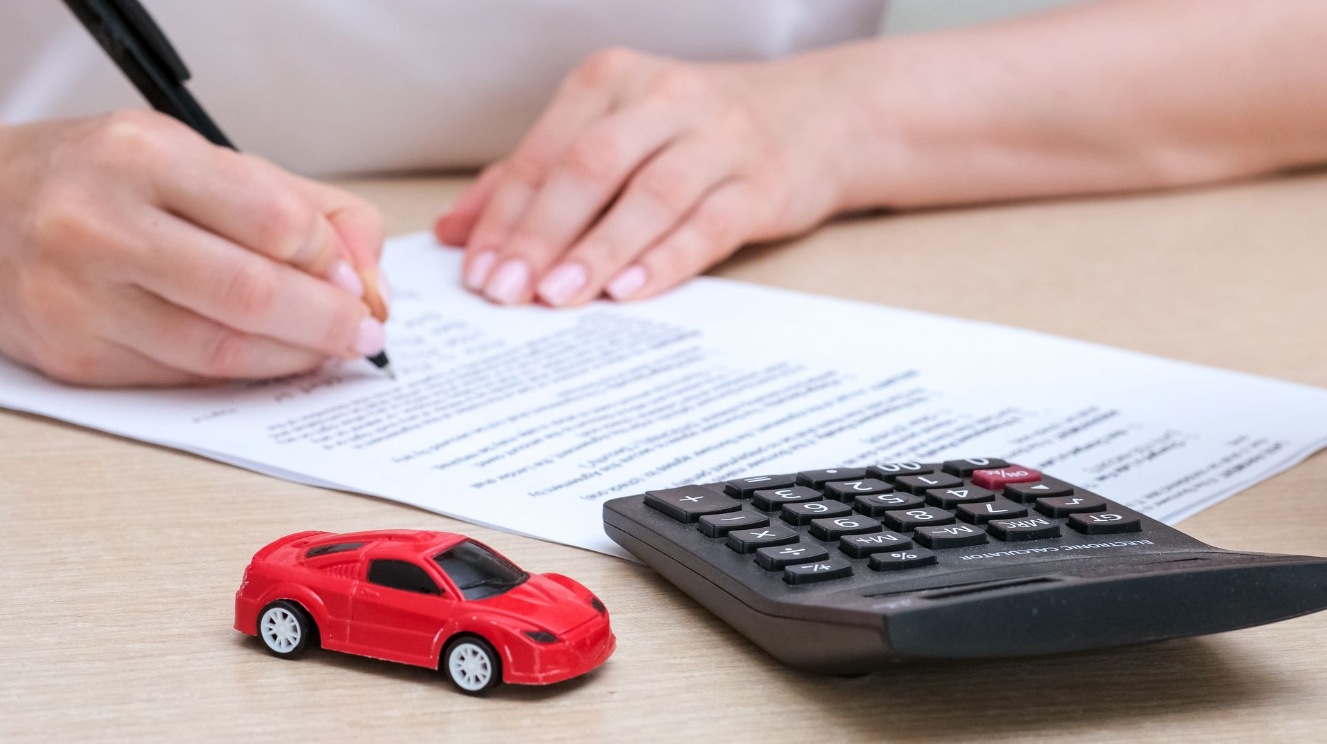 Persona che scrive un documento con una calcolatrice e una macchinina rossa giocattolo su un tavolo.