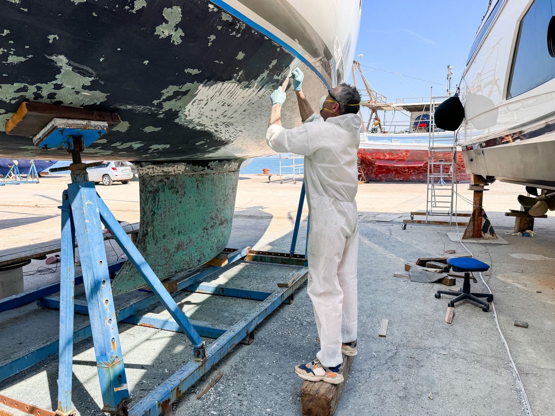 Un uomo in tuta bianca lavora sullo scafo di una barca in un cantiere navale in una giornata di sole.