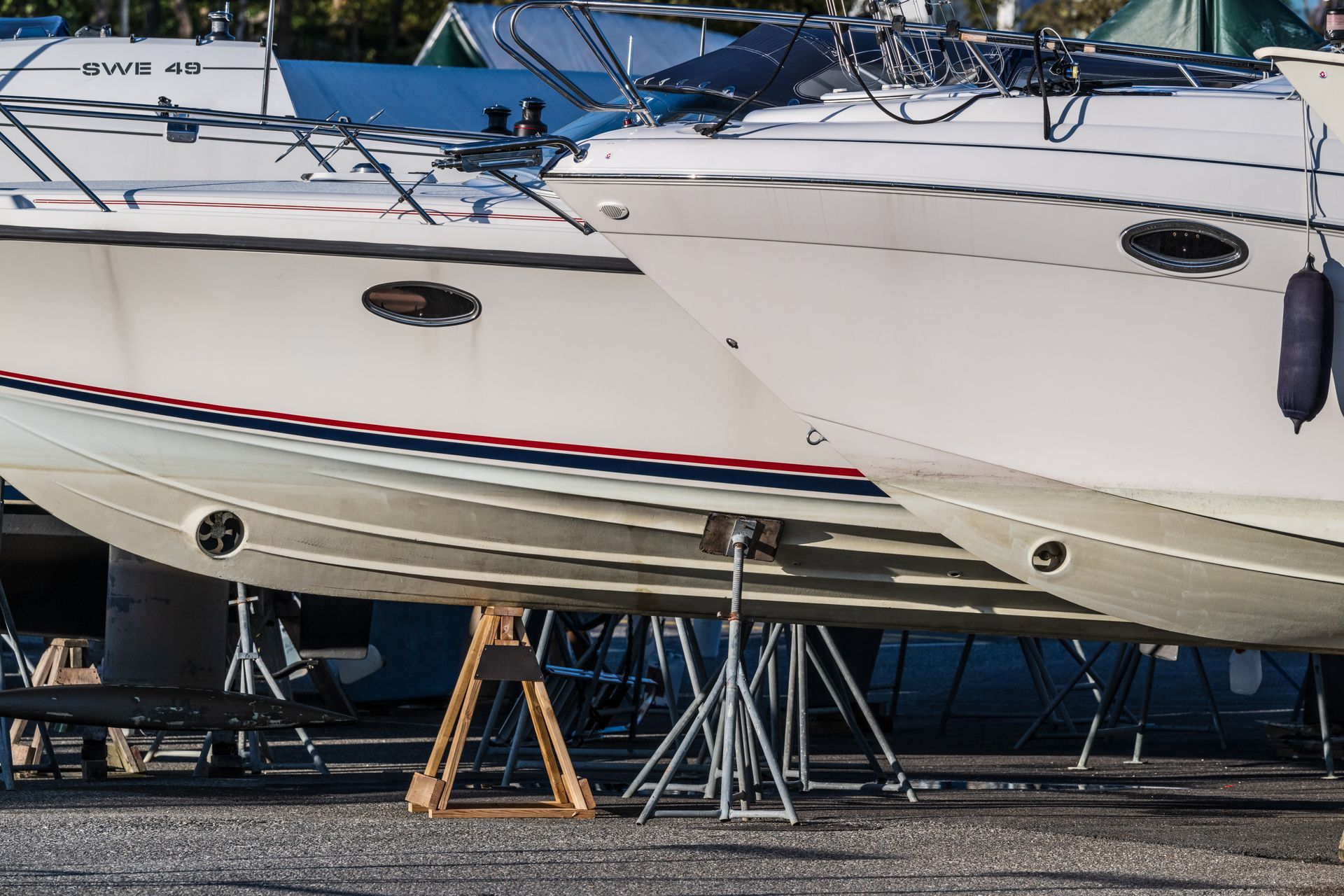 Barche su supporti in un cantiere navale; scafi bianchi con strisce blu e rosse, oblò e barche coperte sullo sfondo.