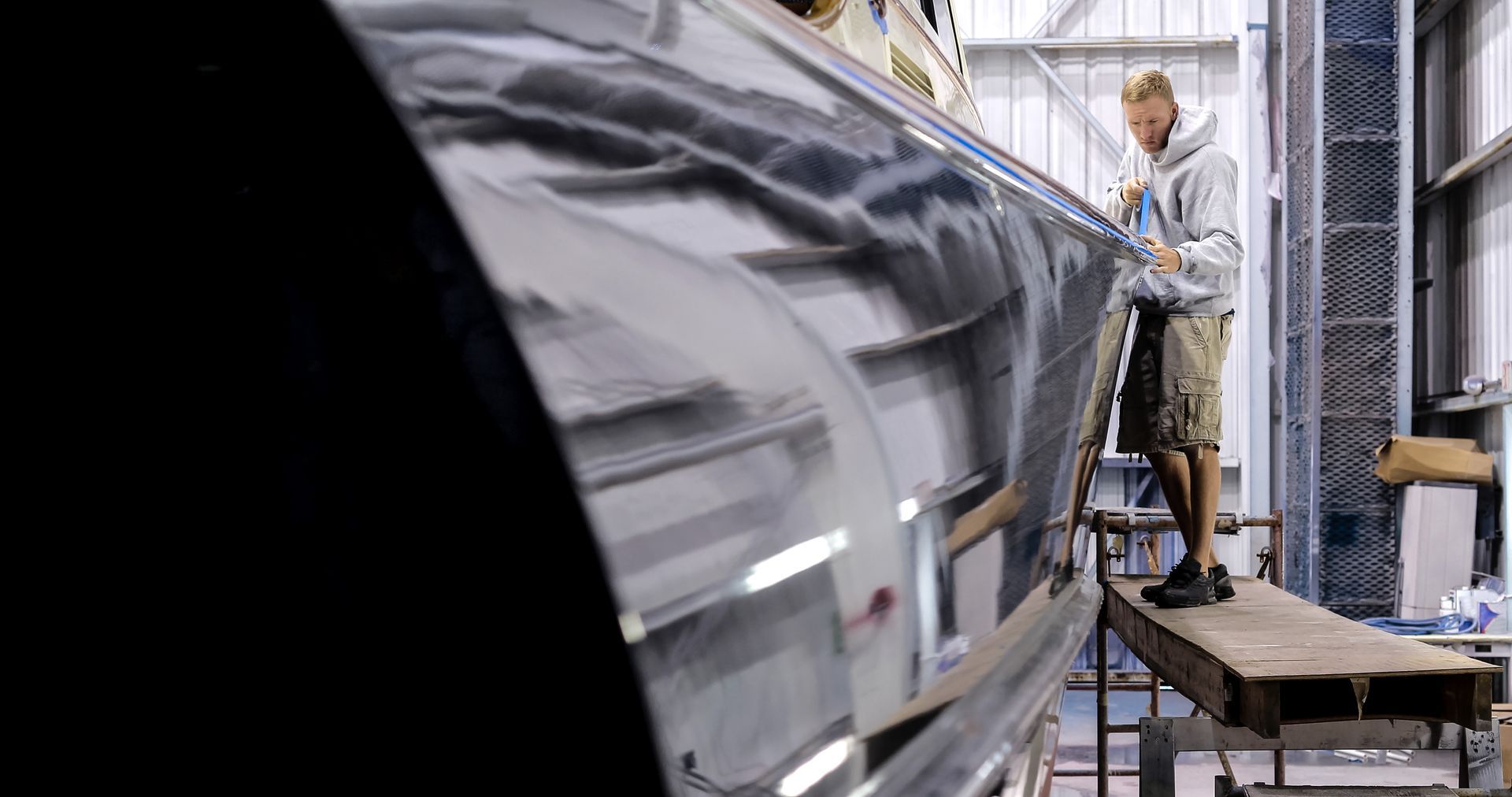 Uomo al lavoro sullo scafo di una barca all'interno di un'officina. Lo scafo nero riflette l'ambiente circostante.