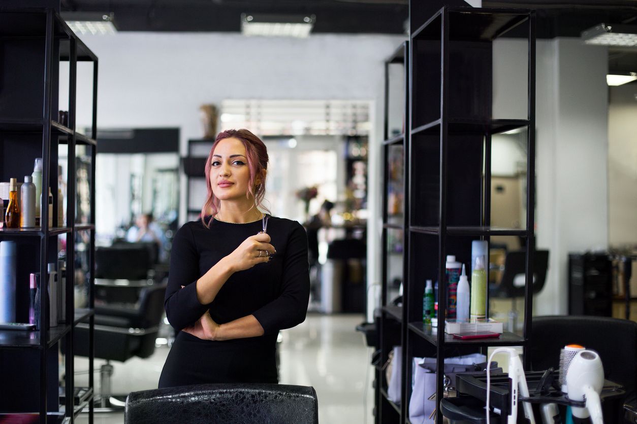 Hair stylist with pink hair in a salon, holding scissors, and smiling.