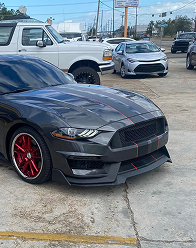A dark gray Ford Mustang with red racing stripes and red wheels parked on a paved lot near other vehicles.