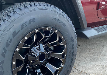 A close-up of a large black and silver off-road wheel mounted on a red Jeep vehicle parked on concrete.