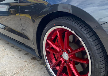 A closeup of a black car's wheel featuring a glossy red multi-spoke rim with a silver outer edge, parked on concrete.