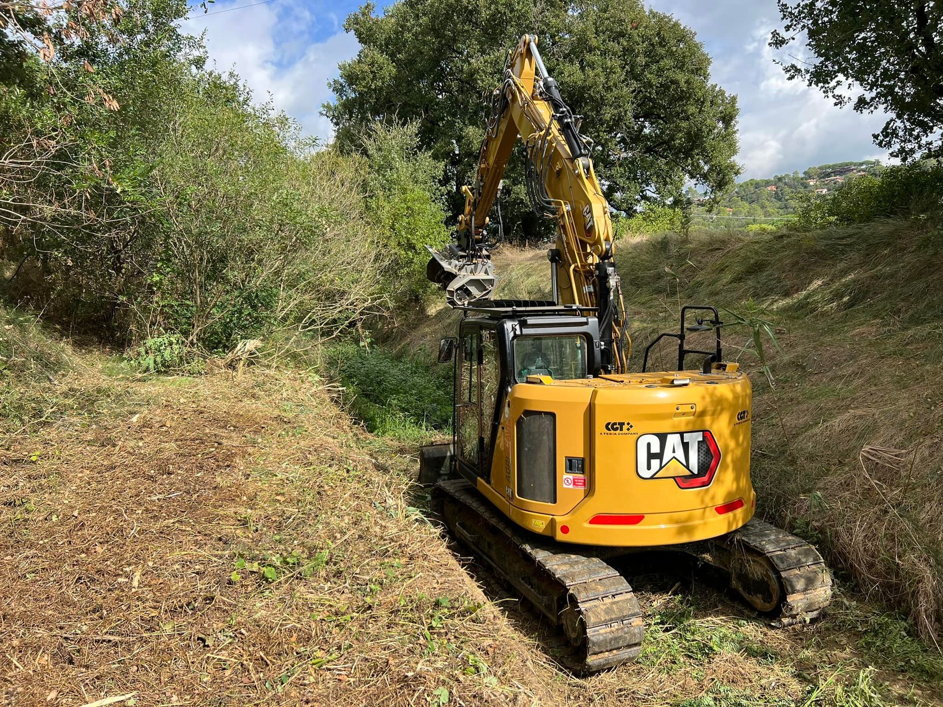 Un escavatore Caterpillar giallo ripulisce la vegetazione vicino a una collina, sotto un cielo parzialmente nuvoloso.