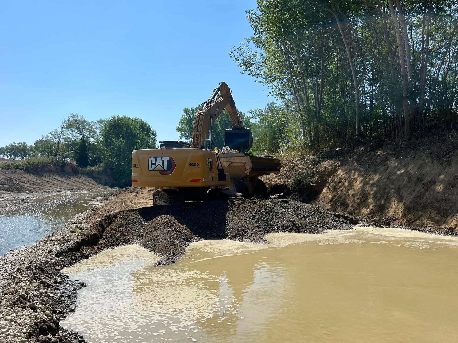 Un escavatore giallo su una riva fangosa raccoglie materiale da uno stagno.