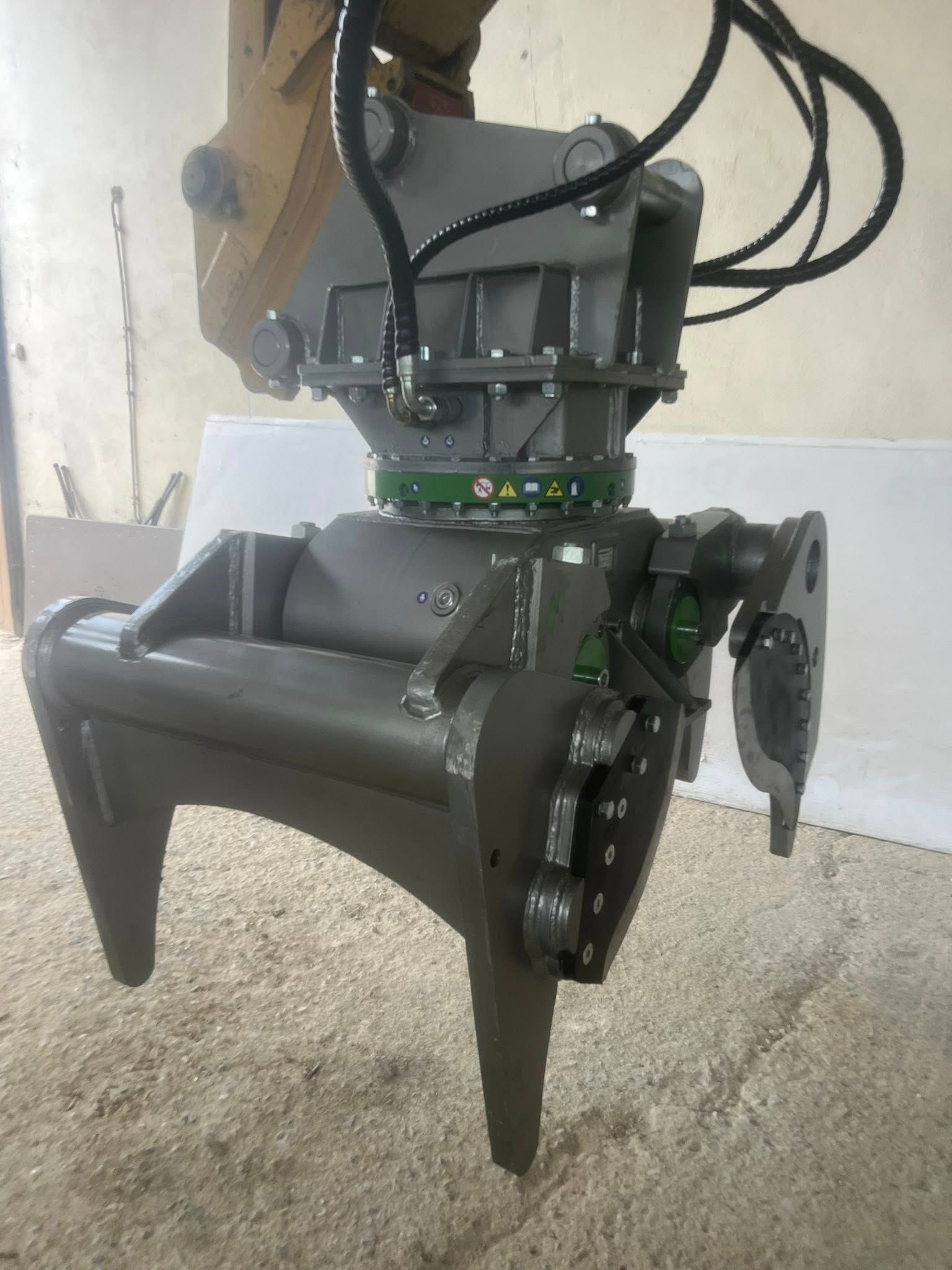 Un grande attacco a pinza grigio per il braccio di un escavatore, appoggiato su una superficie di colore chiaro.