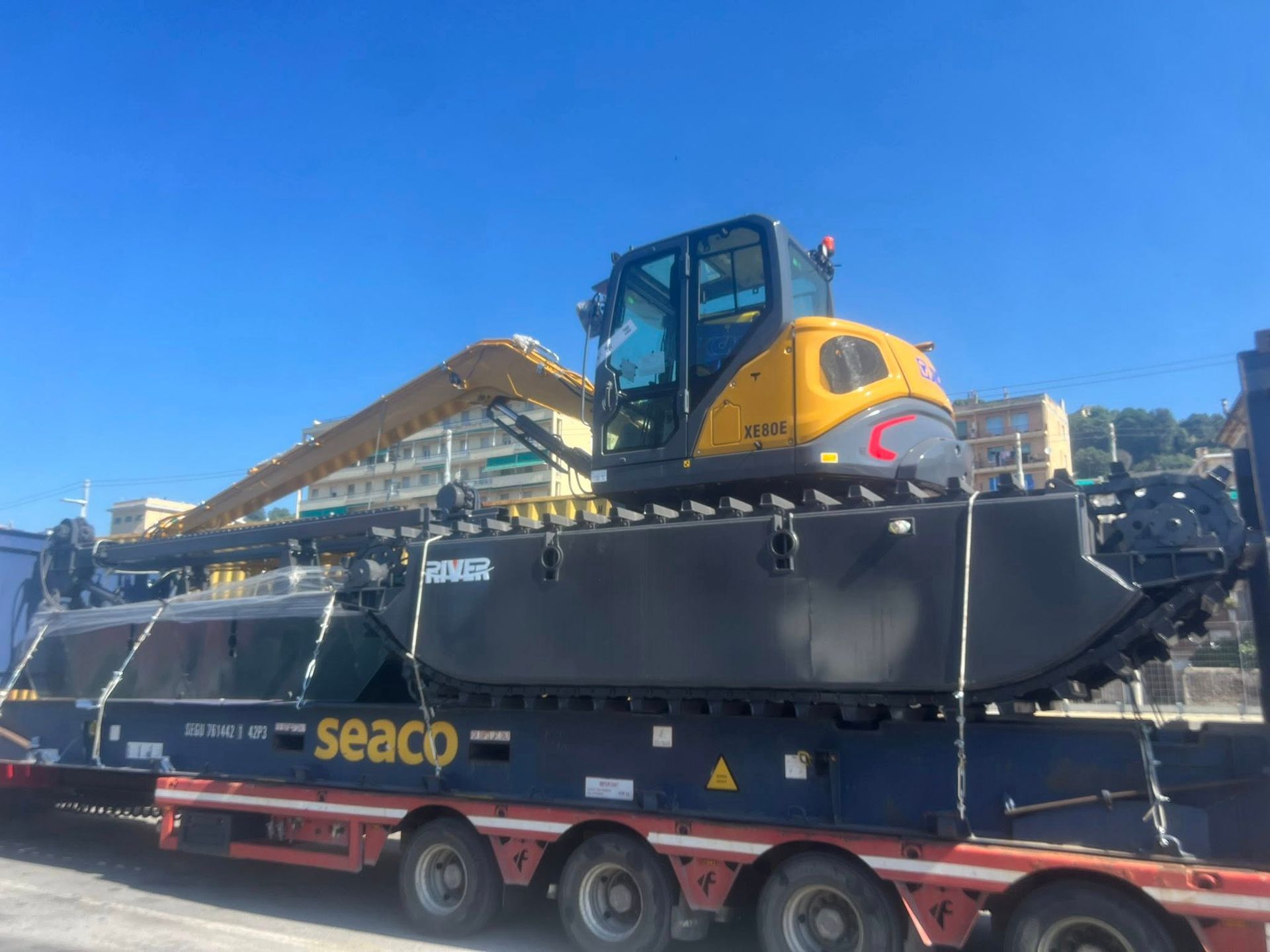Un escavatore anfibio Seaco su un rimorchio, pronto per il trasporto. Cabina gialla, scafo nero. Cielo azzurro e limpido.