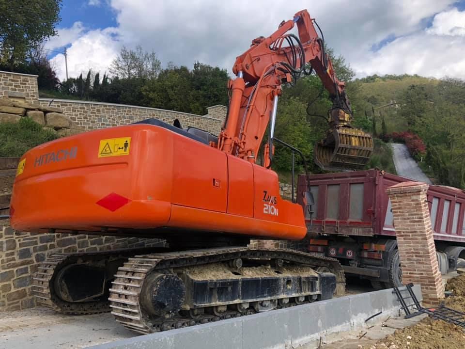Escavatore arancione che carica terra su un autocarro con cassone ribaltabile color marrone; cantiere con muro di pietra e pendio.