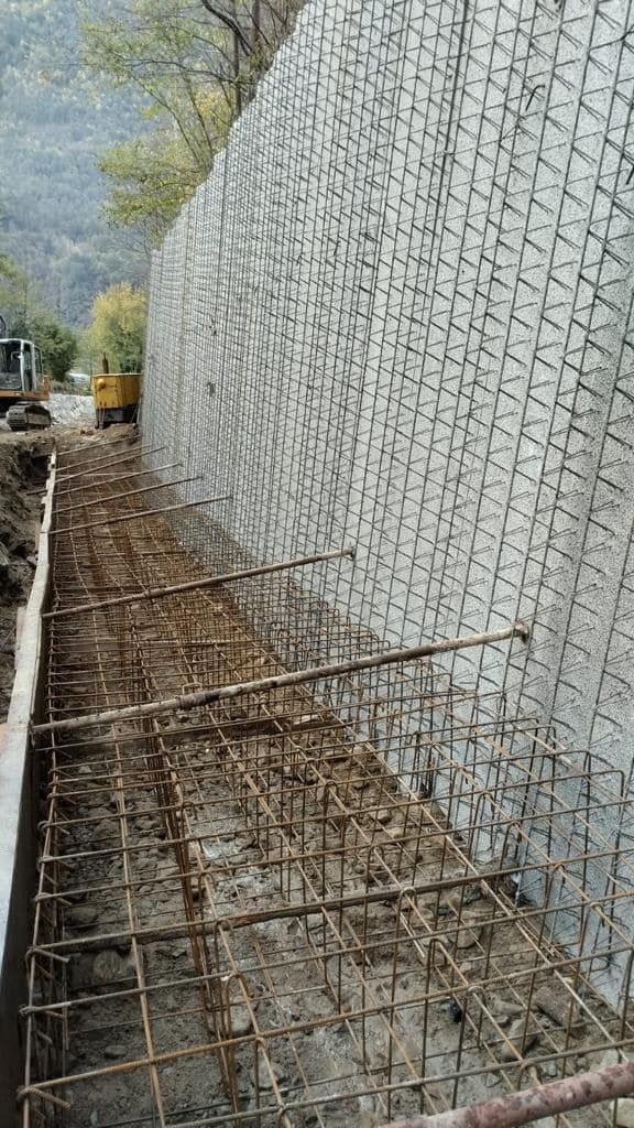 Costruzione di un muro di contenimento con barre di armatura e rete metallica a vista in un contesto collinare.