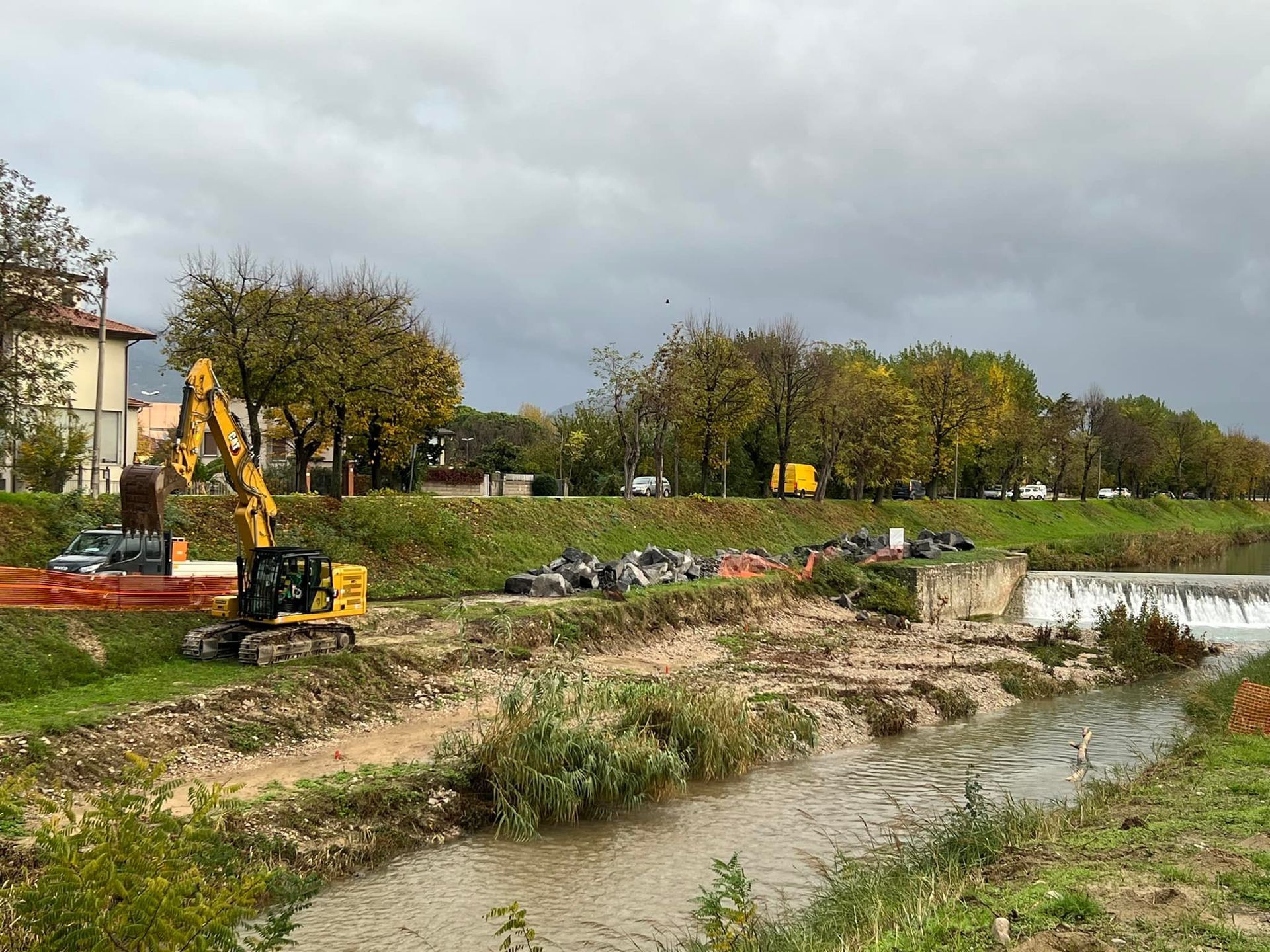 Lavori in corso lungo la riva di un fiume, con un escavatore, detriti e una piccola cascata sotto un cielo nuvoloso.