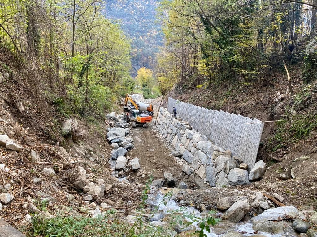 Cantiere in una zona boscosa con escavatori, pareti rocciose e un ruscello.